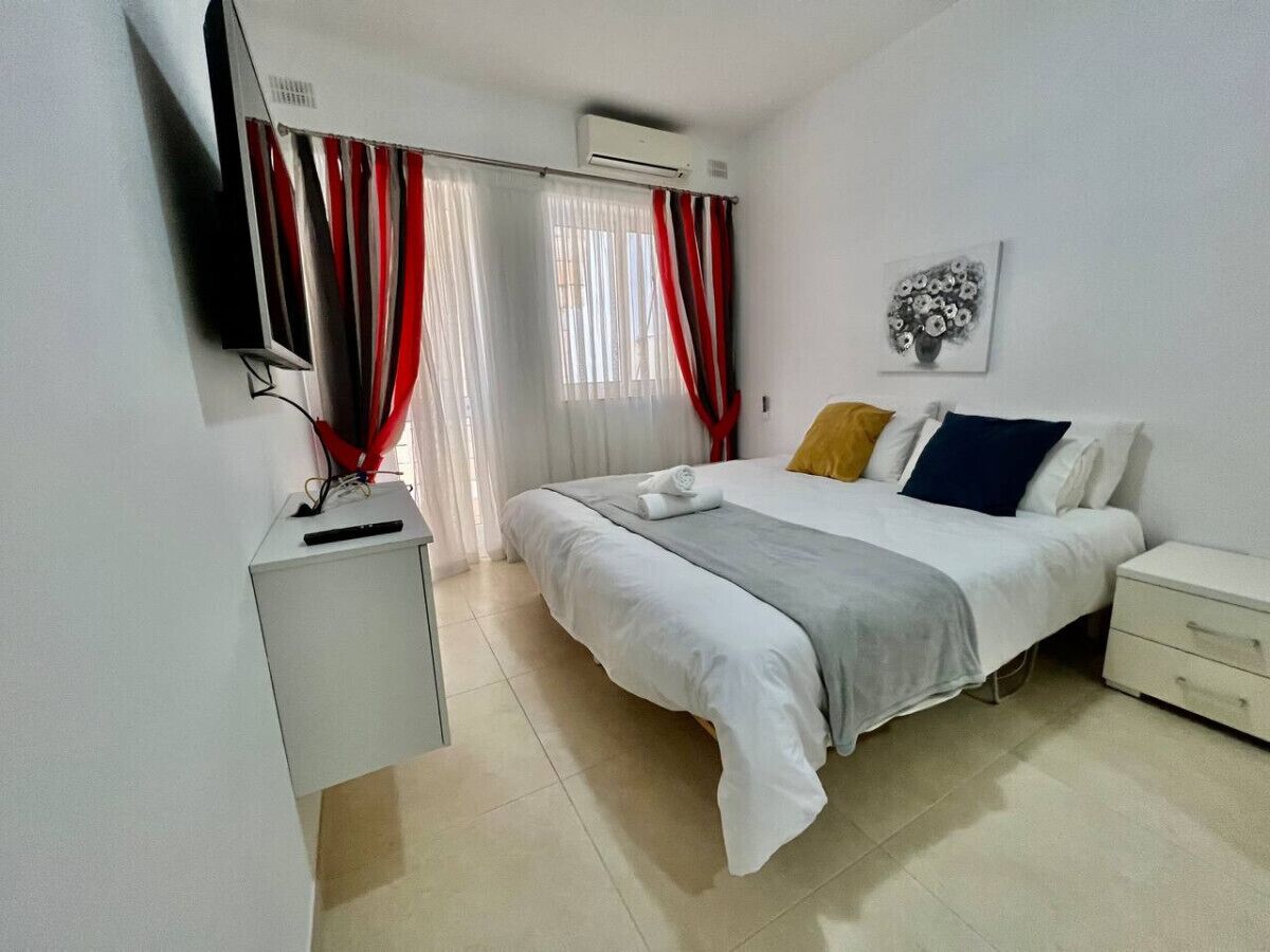 Een slaapkamer met een bed, nachtkastje, tv aan de muur en een raam met gordijnen.