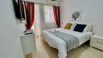 Een slaapkamer met een bed, nachtkastje, tv aan de muur en een raam met gordijnen.