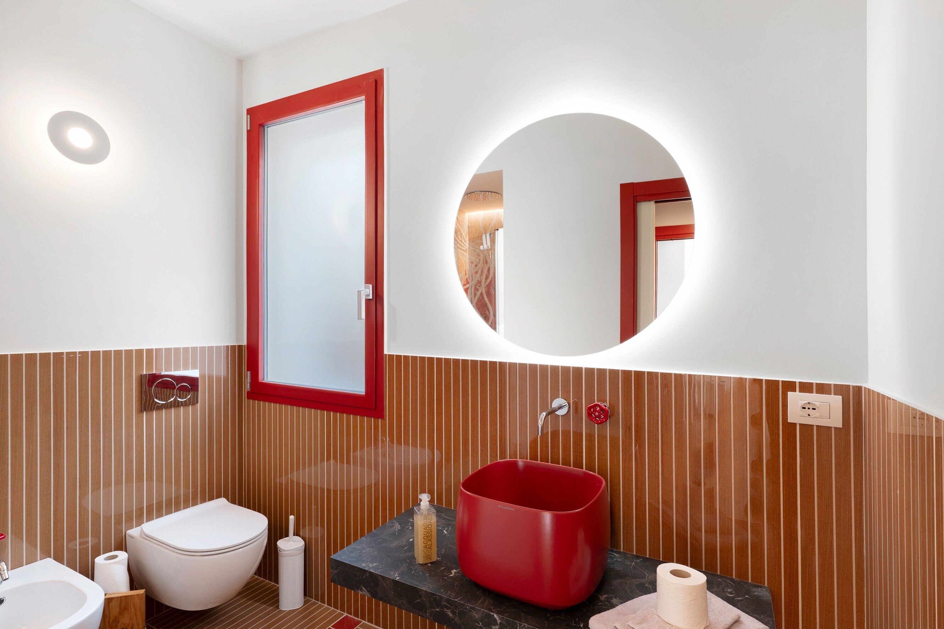 Ein modernes Badezimmer mit Toilette in Weiß, einem roten Waschbecken, einem Spiegel und Holzvertäfelung mit Streifen.