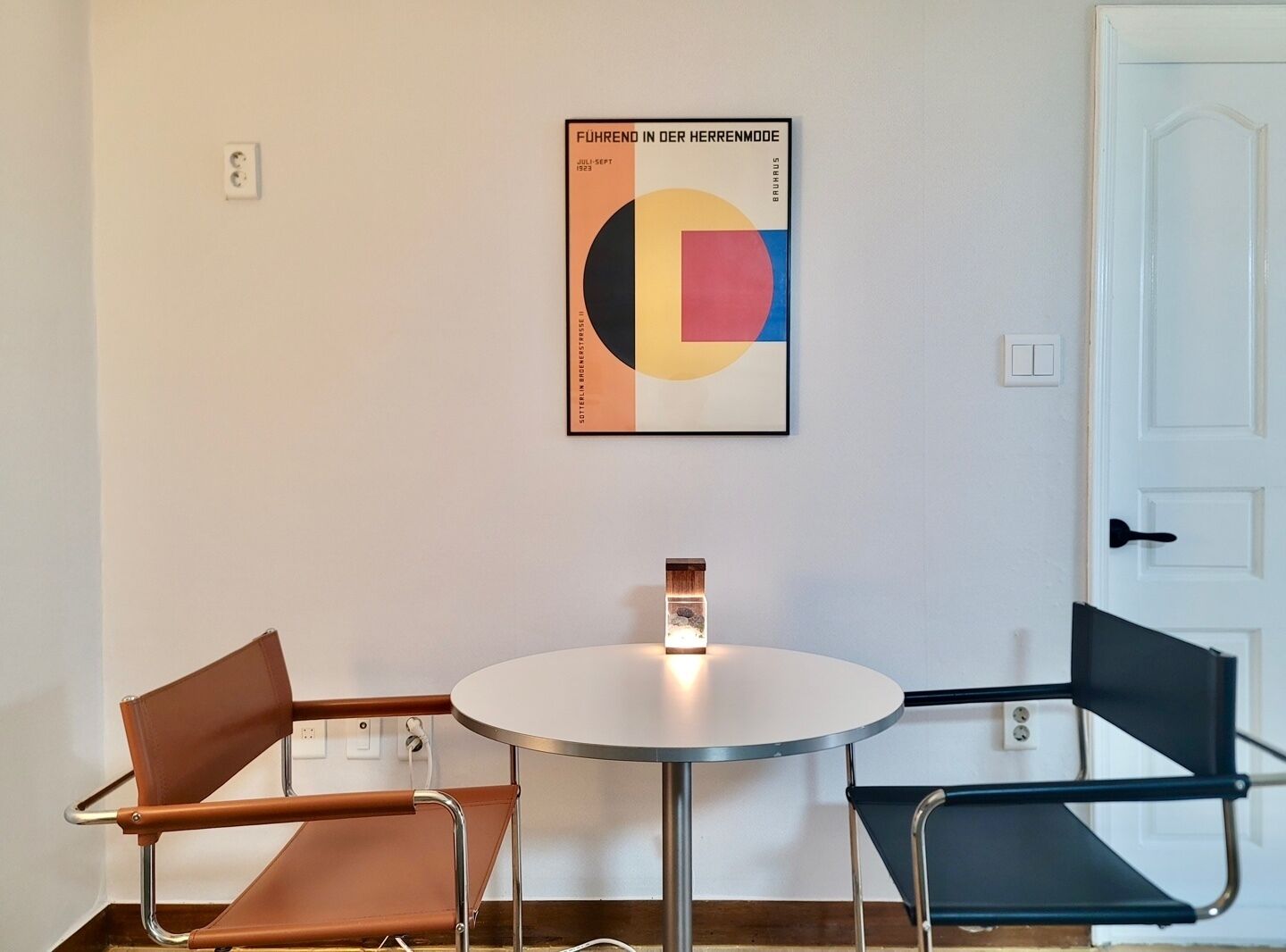 A room with a round table and two chairs, one brown and one black. A colorful poster is on the wall.