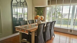 Un coin repas avec une table en bois, des chaises en osier, un miroir et une grande fenêtre avec des stores.