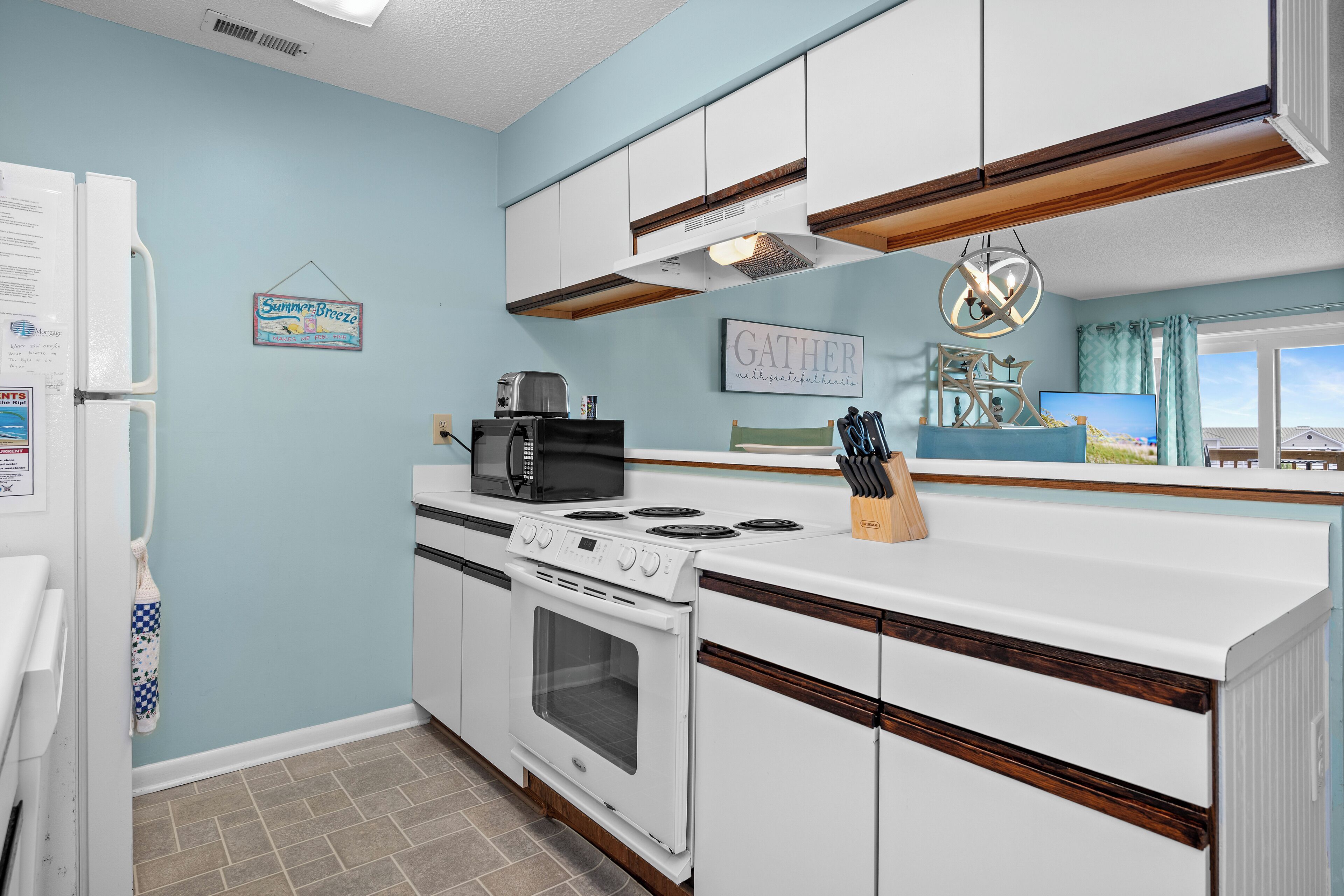 Une cuisine avec un four, un four à micro-ondes et une cuisinière, tous de couleur blanche. Un panneau suspendu porte l’inscription « Summer Breeze », et un tableau encadré affiche le mot « GATHER ».