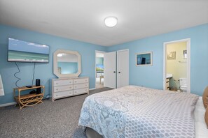 A bedroom with a bed, dresser, TV, and a view of the beach through an open door. - Oceanview 2BR Condo w/ Pool, Easy Beach Access, Private Deck & Relaxing Escape (Emerald Isle)