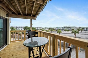 A wooden balcony with a glass table and chairs, overlooking a parking area and buildings. - Oceanview 2BR Condo w/ Pool, Easy Beach Access, Private Deck & Relaxing Escape (Emerald Isle)