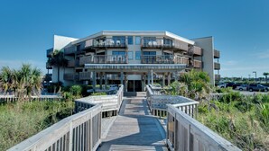 Un hotel de varios pisos con balcones, un paseo marítimo de madera que lleva a la entrada y un cielo azul despejado.