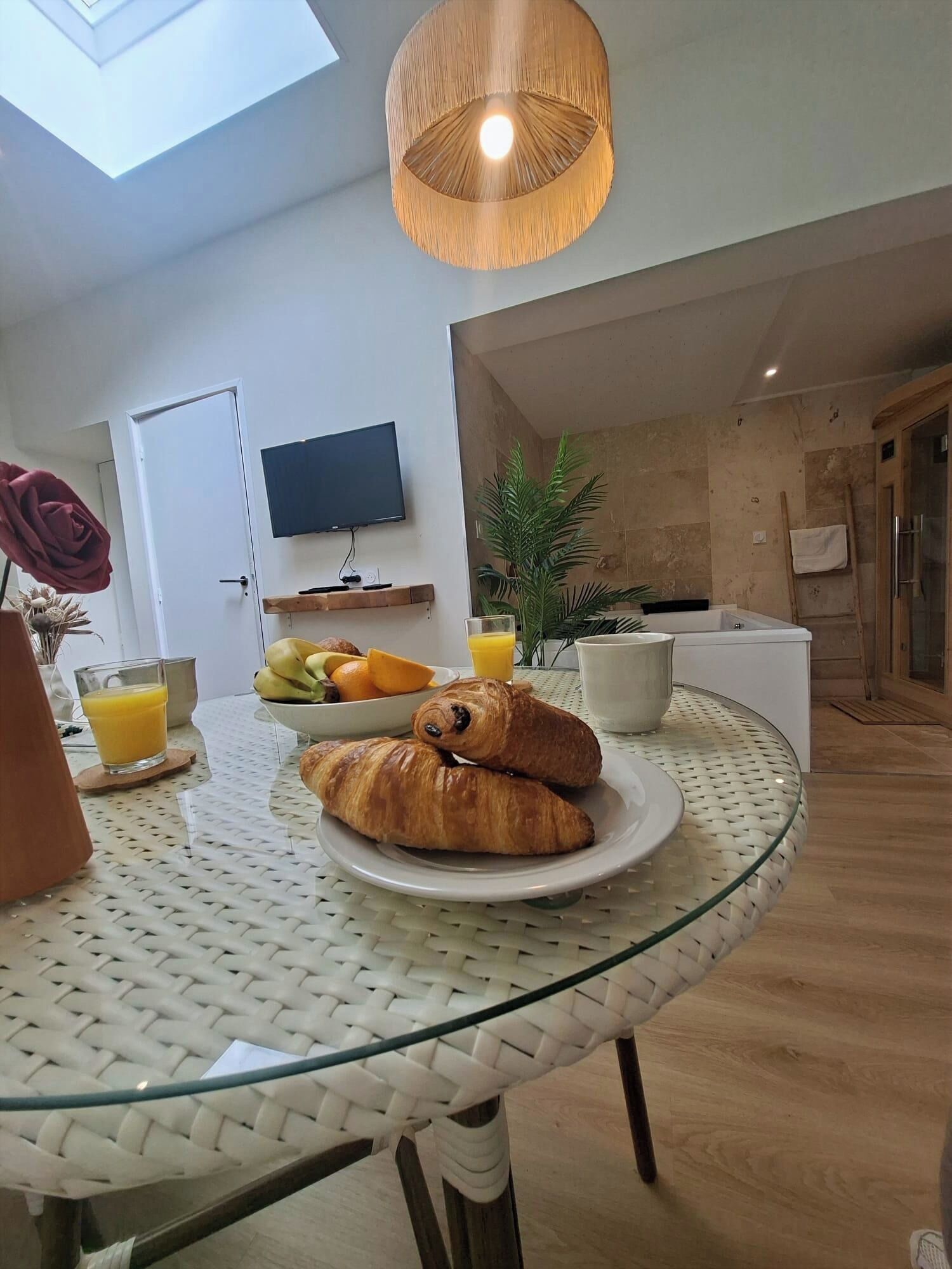 A breakfast setup with croissants, fruit, and juice on a glass table, a TV on the wall, and a modern kitchen in the background.