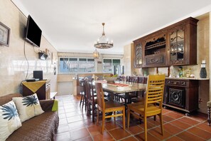 Un coin repas avec une table ronde, des chaises, un canapé agrémenté de coussins décoratifs, un téléviseur fixé au mur, et une cuisine avec des meubles en bois et un lustre.