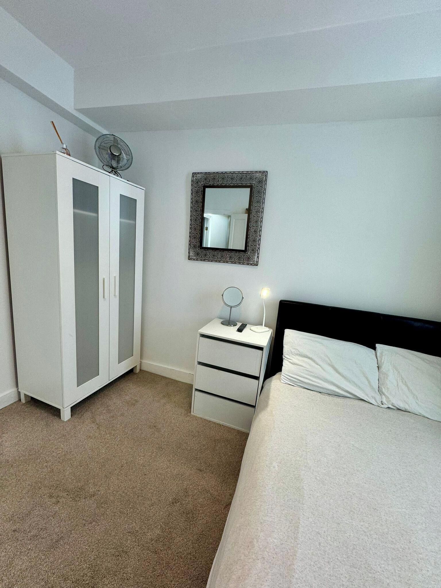 Une chambre à coucher comprenant un lit, une table de chevet avec une lampe, un miroir, une armoire et un ventilateur de plafond.