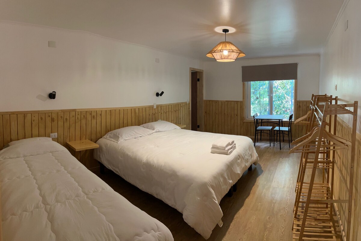 Ein Zimmer mit zwei Betten, einem kleinen Tisch, einem Stuhl, einer Pendelleuchte und einem Fenster mit Blick auf Bäume.