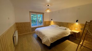 Une chambre avec un plancher en bois, un lit avec une literie blanche, une fenêtre donnant sur des arbres et un luminaire au plafond.