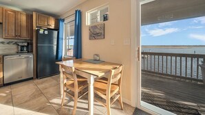 A kitchen with wooden cabinets, a black refrigerator, and a dining table with chairs. There is a view of a wooden deck and water through a glass door.