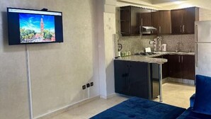 Ein Wohnzimmer mit an der Wand montiertem Fernseher, einem blauen Sofa und einer Küchenzeile mit dunklen Schränken und einer Edelstahl-Dunstabzugshaube.