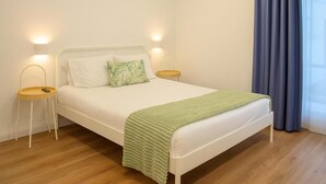 A neatly made bed with white bedding and a green throw, two bedside tables with lamps, and a large window with blue curtains. - Magnolia Suites & Stays (Porto)