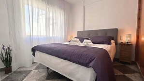 Een net opgemaakt bed met een paars-witte dekbedovertrek, een bedlampje en een klein houten nachtkastje met een lamp.