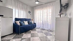 Een kamer met een blauwe bank, witte gordijnen, een plafondventilator en een betegelde vloer.