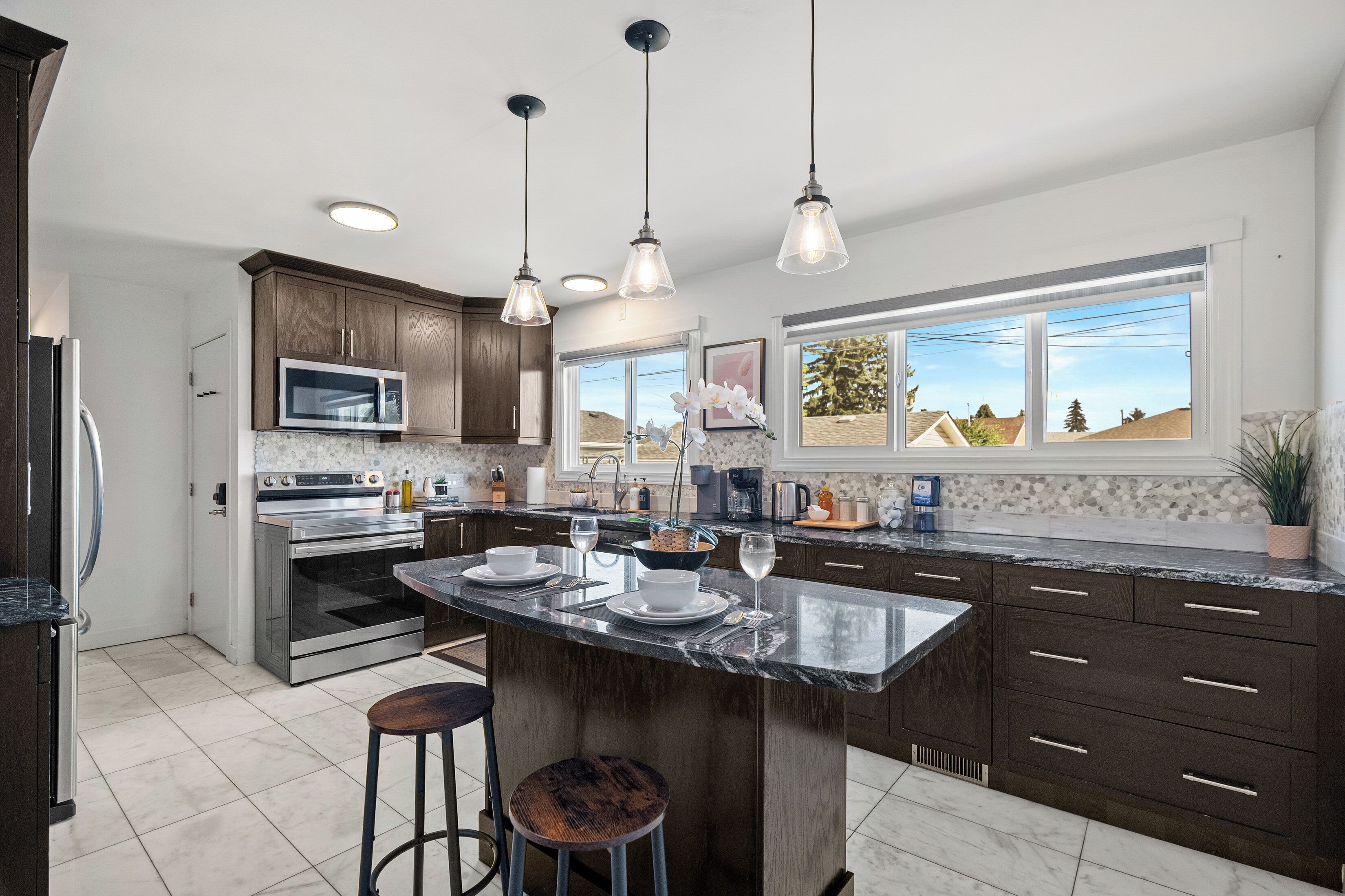 A modern kitchen with dark wood cabinets, stainless steel appliances, a large island with stools, pendant lighting, and a view of the outdoors through large windows.