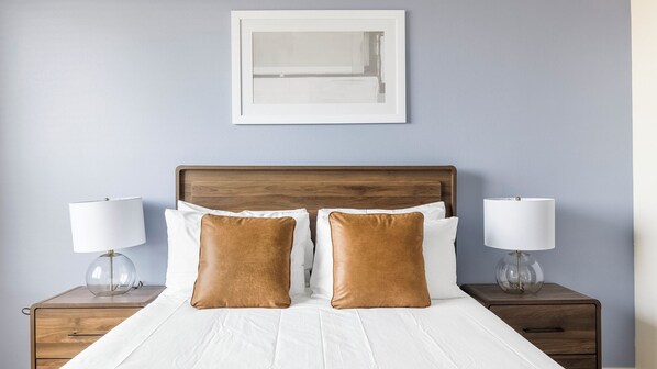 A neatly made bed with white linens and two brown pillows, flanked by two bedside tables with lamps, against a light blue wall with a framed picture above. - Landing Furnished Apartments - Plano (Plano)
