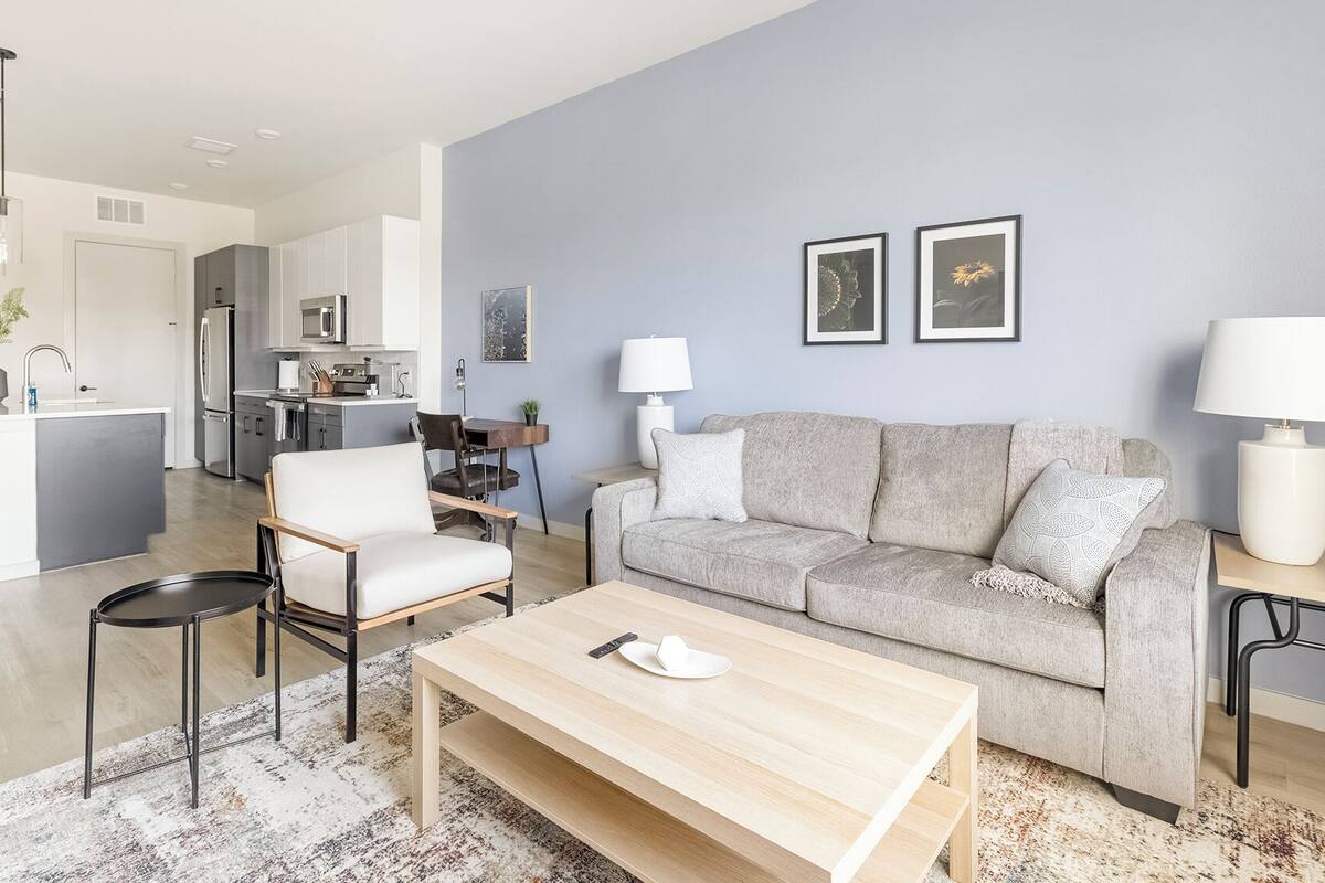 A modern living room with a grey sofa, a wooden coffee table, and a dining area with a kitchen in the background.
