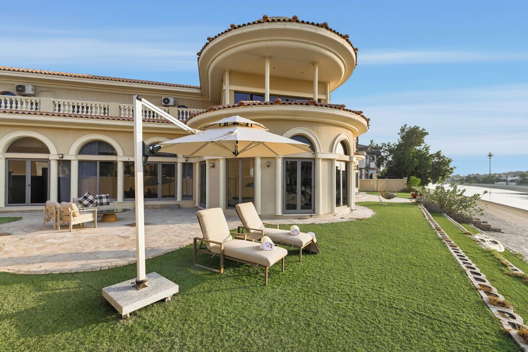 A two-story house with a circular balcony, outdoor seating area with chairs and an umbrella, and a well-maintained lawn.