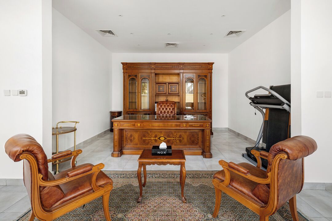 A spacious room with a large wooden desk, two leather armchairs, a treadmill, and a patterned rug.