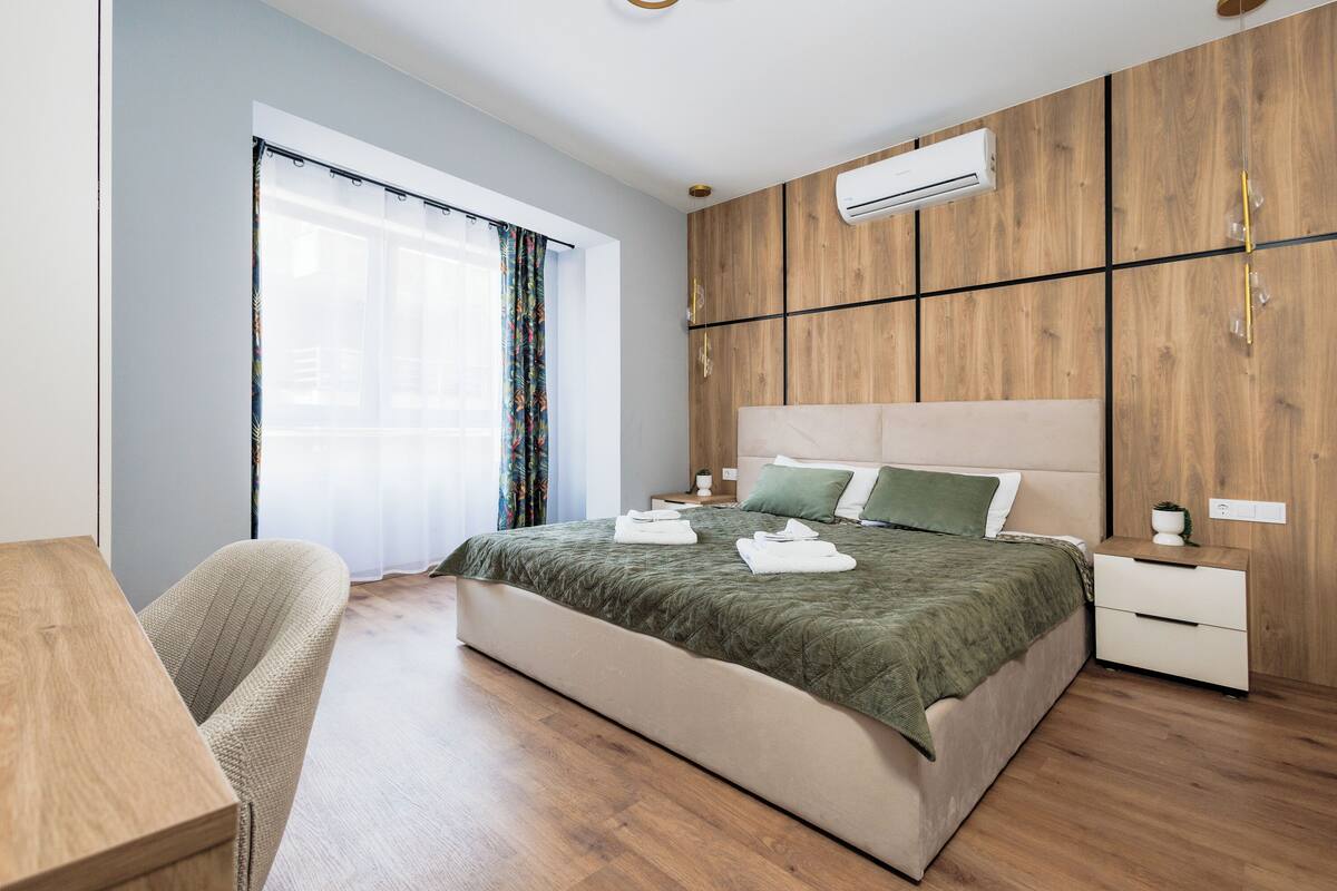 Egy modern hálószoba, melyben van egy fafejű ágy, egy zöld takaróval borított ágy, egy éjjeliszemély és egy függönnyel ellátott ablak.