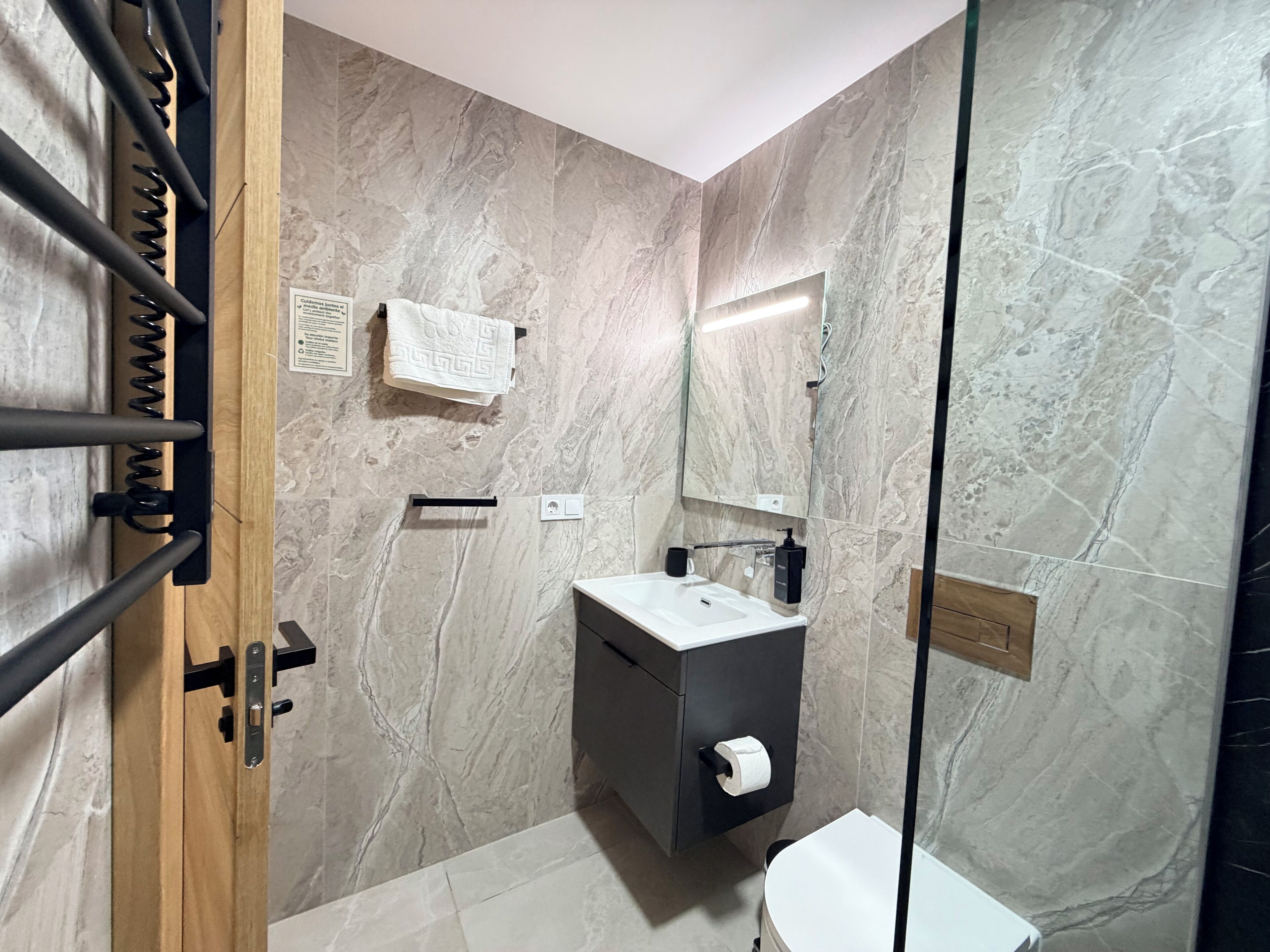 A modern bathroom with a marble wall, a wooden door, a black cabinet with a toilet paper holder, and a glass shower enclosure.