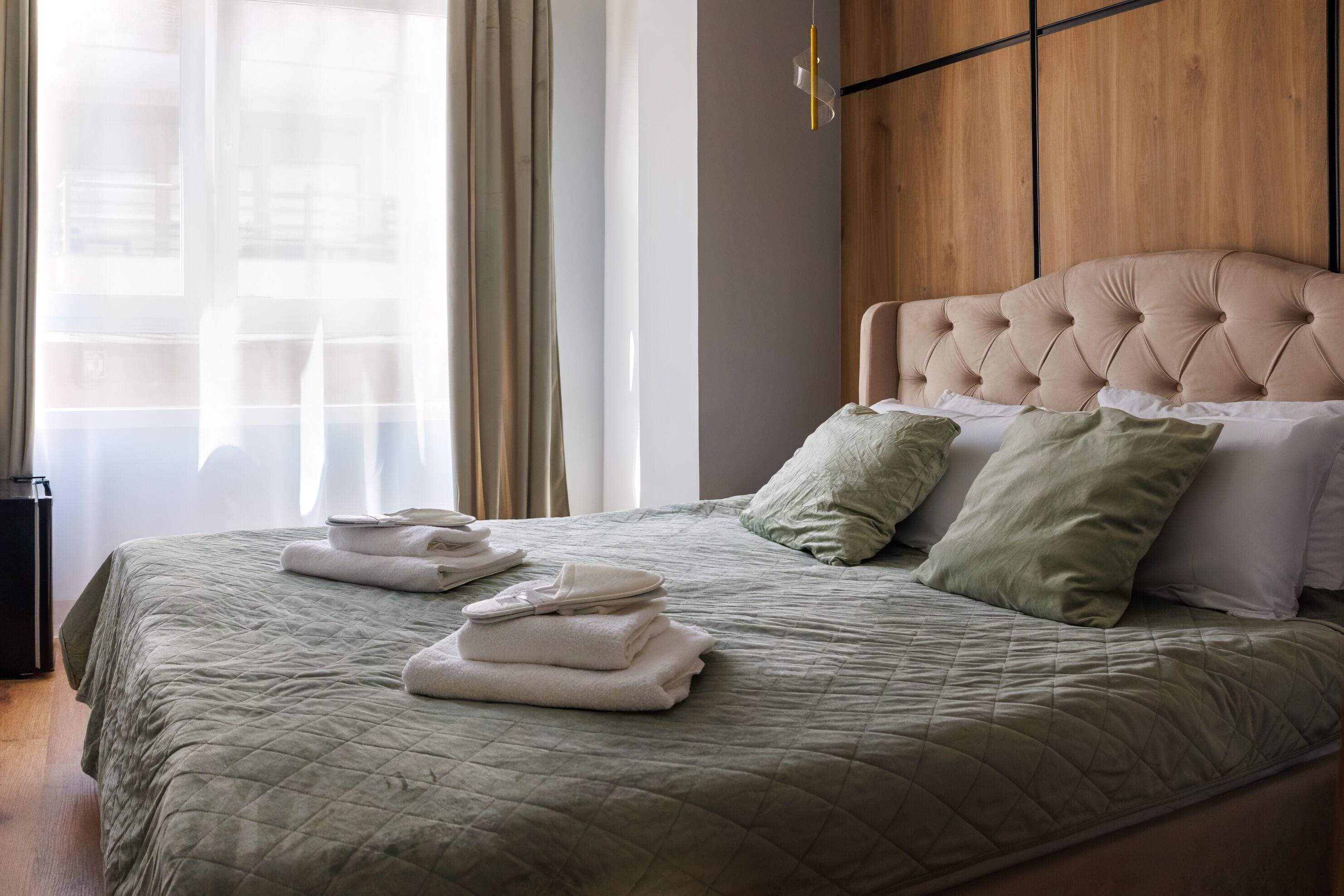 A neatly made bed with a quilted green bedspread, a tufted headboard, and a stack of folded white towels.