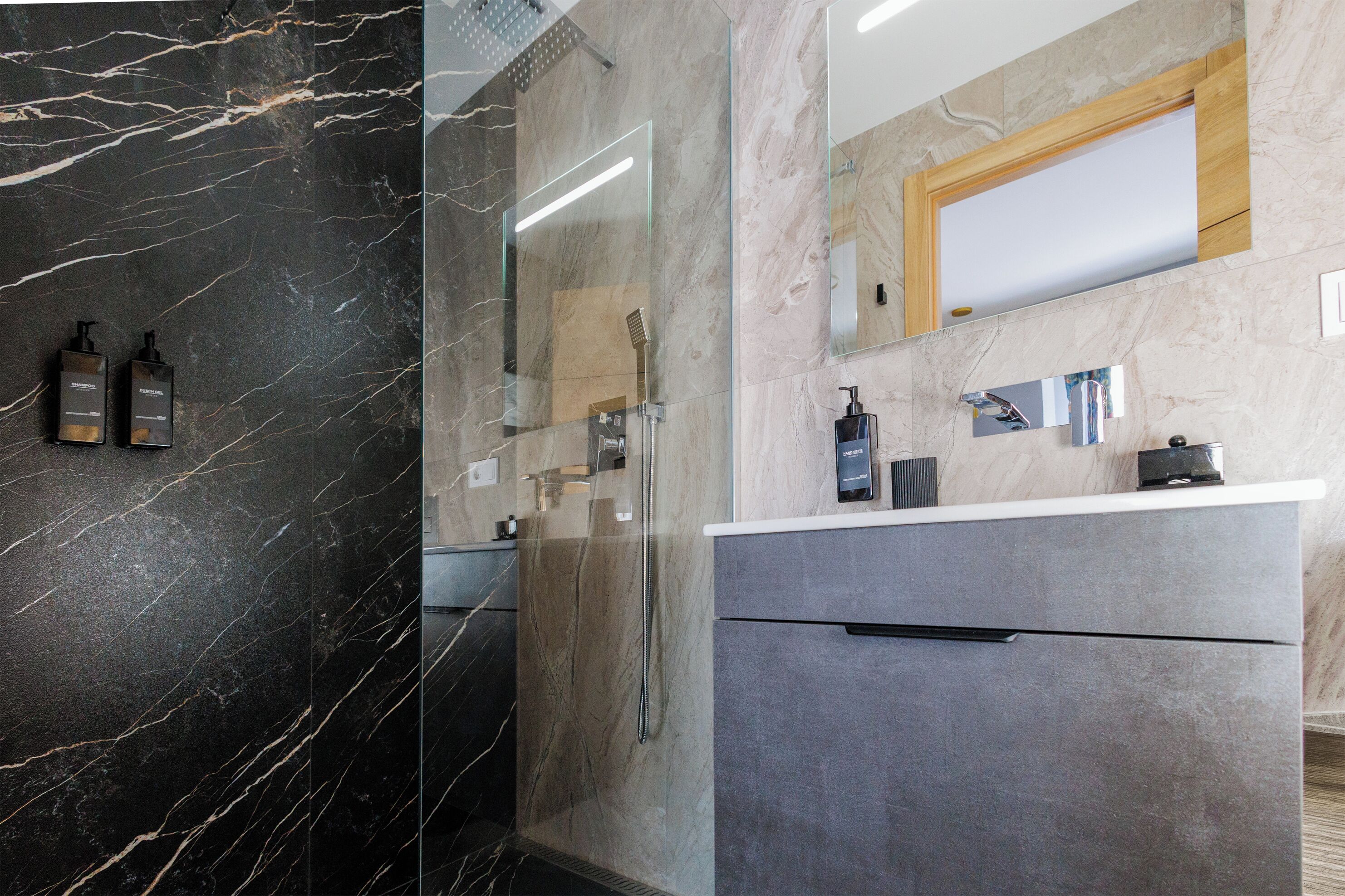 Een badkamer met een marmeren wand, een douche, een wastafel met spiegel en twee flessen aan de wand.