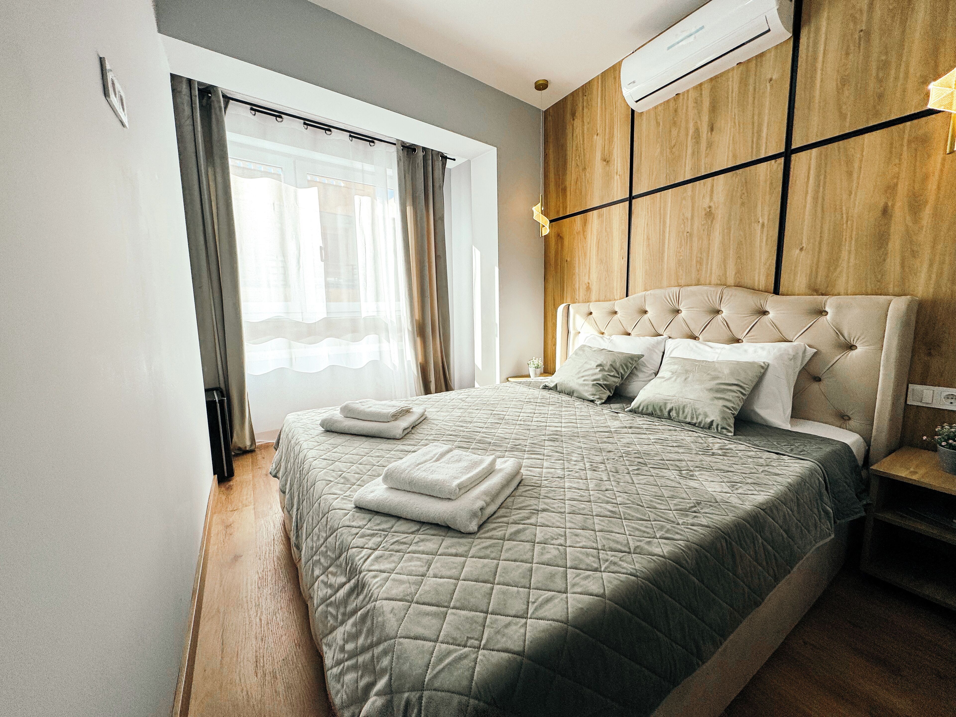A modern bedroom with a large bed, a wooden headboard, and a window with sheer curtains.