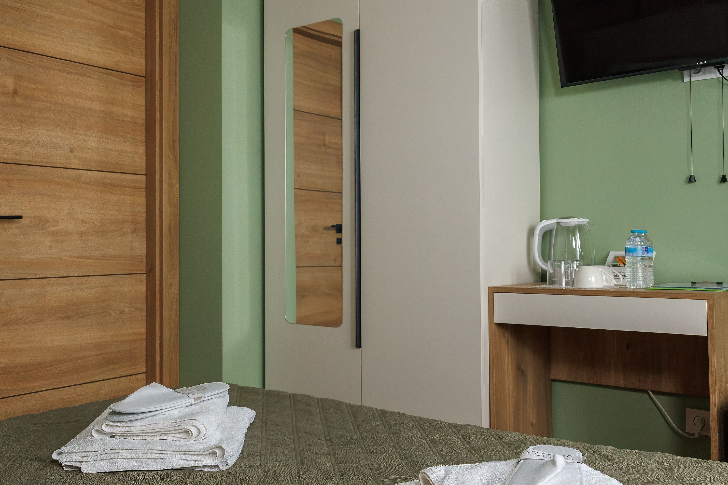 A hotel room with a bed, a wooden wardrobe, a mirror, a desk with a kettle, and a television.