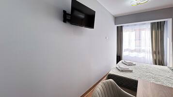 Una habitación de hotel moderna con una televisión de pantalla plana montada en la pared, una cama con cubrecama acolchado, una mesita de noche de madera y una ventana con cortinas.