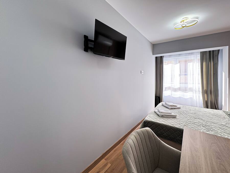 Una habitación de hotel moderna con una televisión de pantalla plana montada en la pared, una cama con cubrecama acolchado, una mesita de noche de madera y una ventana con cortinas.
