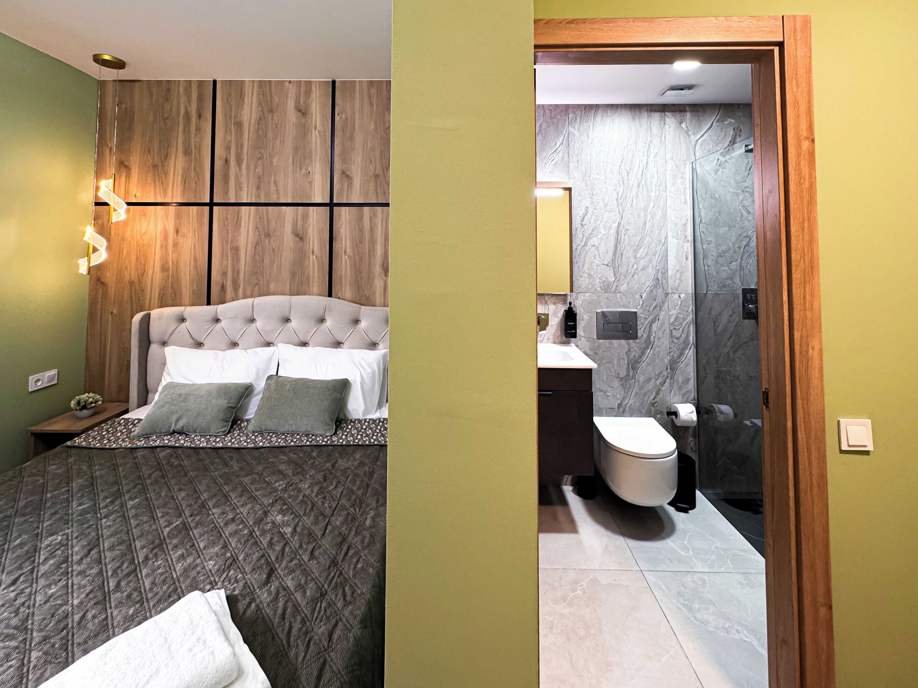 Un dormitorio con cabecera capitonada, cama con ropa de cama estampada y lámparas de noche. El baño tiene un lavamanos moderno, inodoro y paredes de mármol.