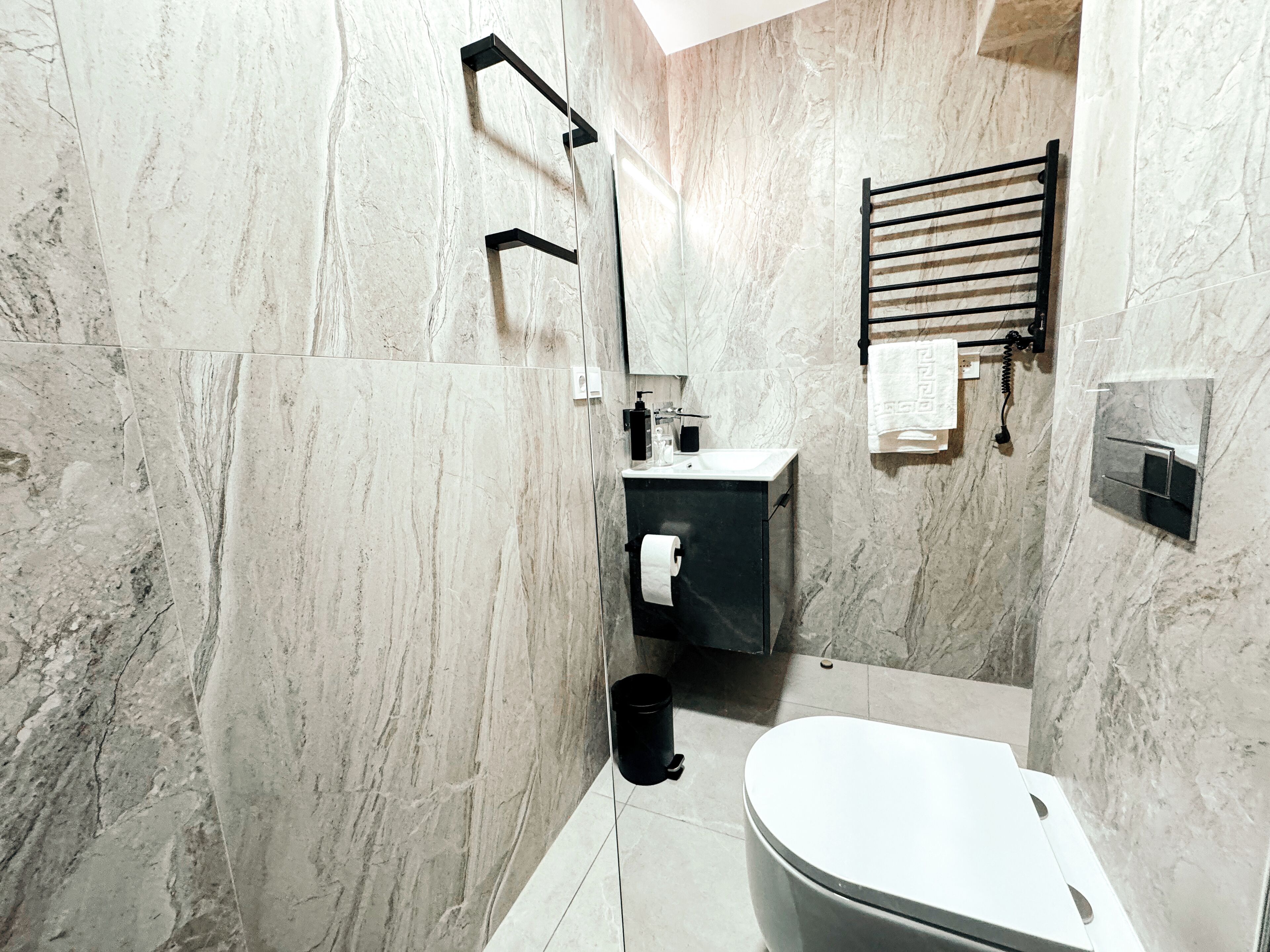 Une salle de bain moderne avec un mur en marbre, un porte-serviettes noir, des toilettes et un lavabo avec un miroir.