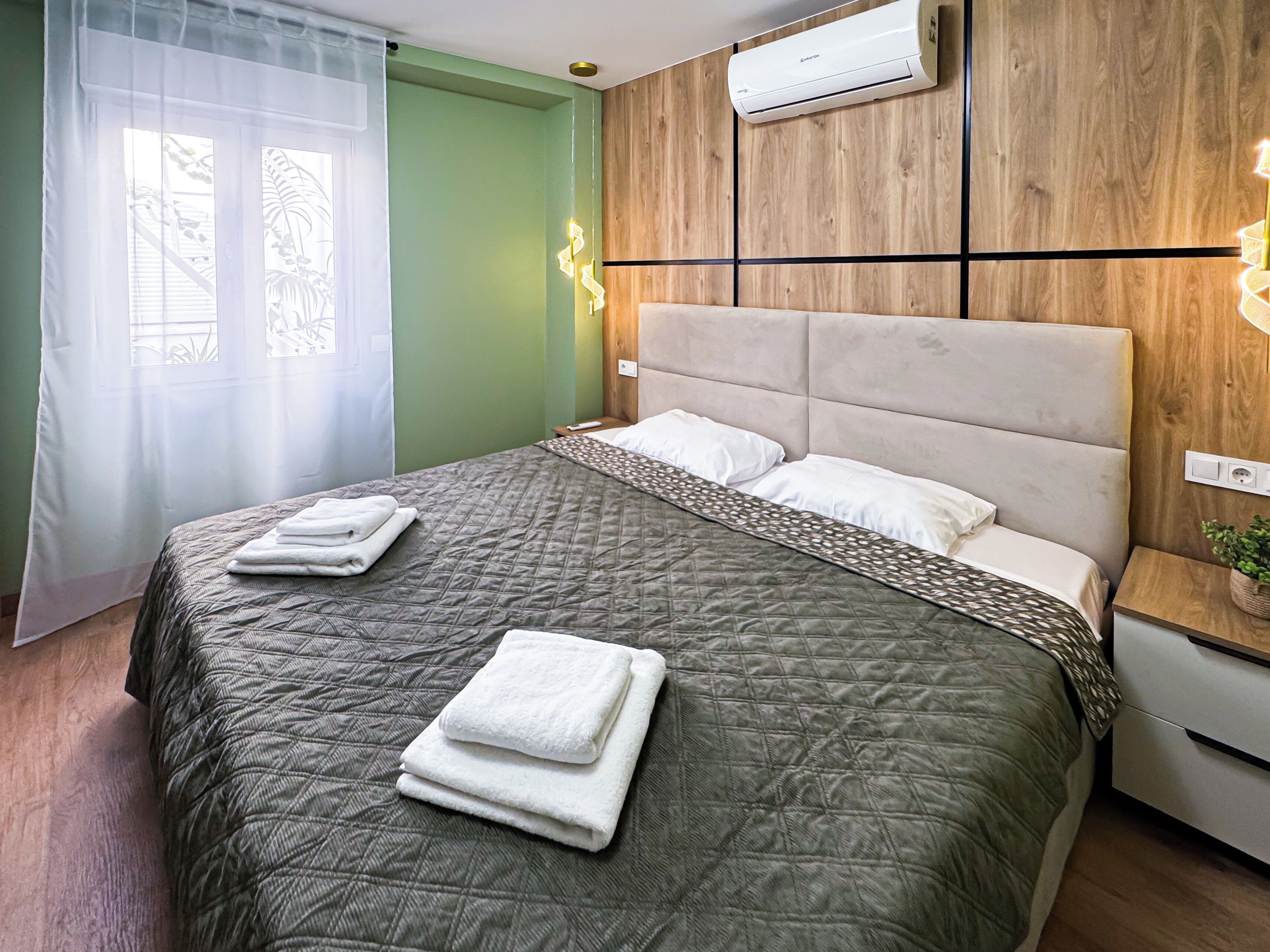 A neatly made bed with a quilted bedspread and folded towels, a wooden panel wall, a wall-mounted air conditioner, and a window with sheer curtains.