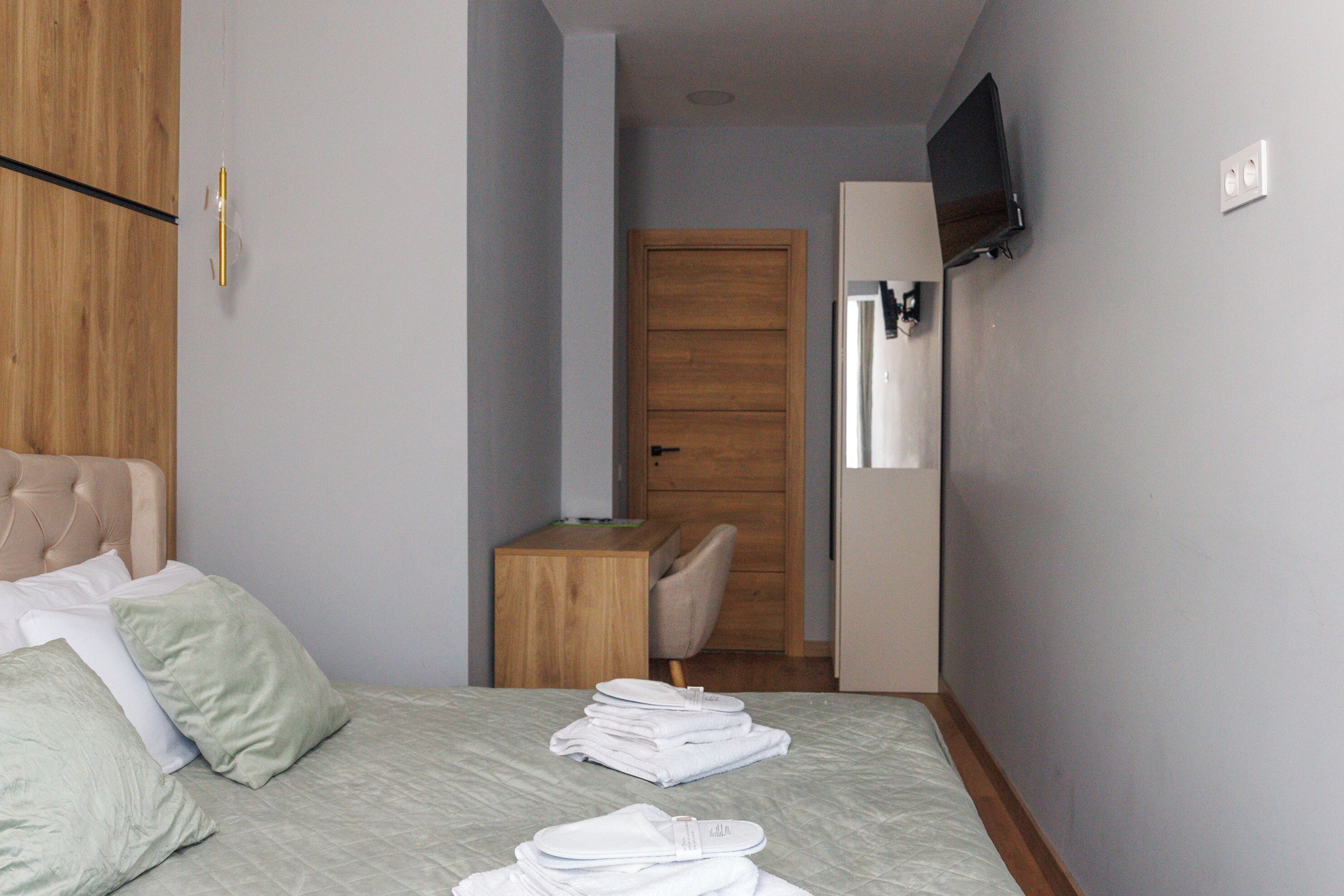 A bedroom with a bed, pillows, a wooden nightstand, a chair, a desk, a television, and a wardrobe.