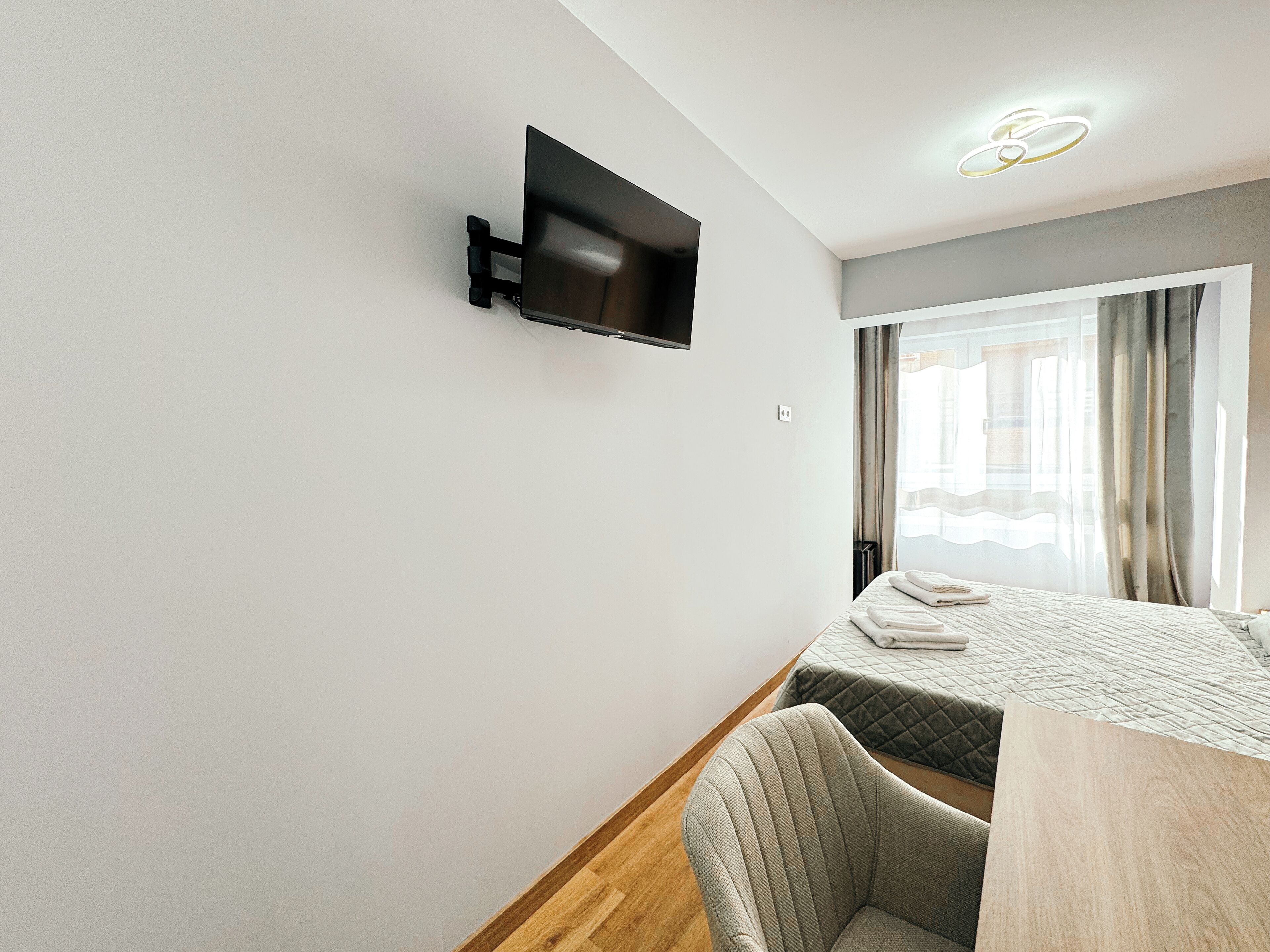 Ein Hotelzimmer mit einem Bett, einem Sessel, einem an der Wand befestigten Fernseher und einem Fenster mit Vorhängen.