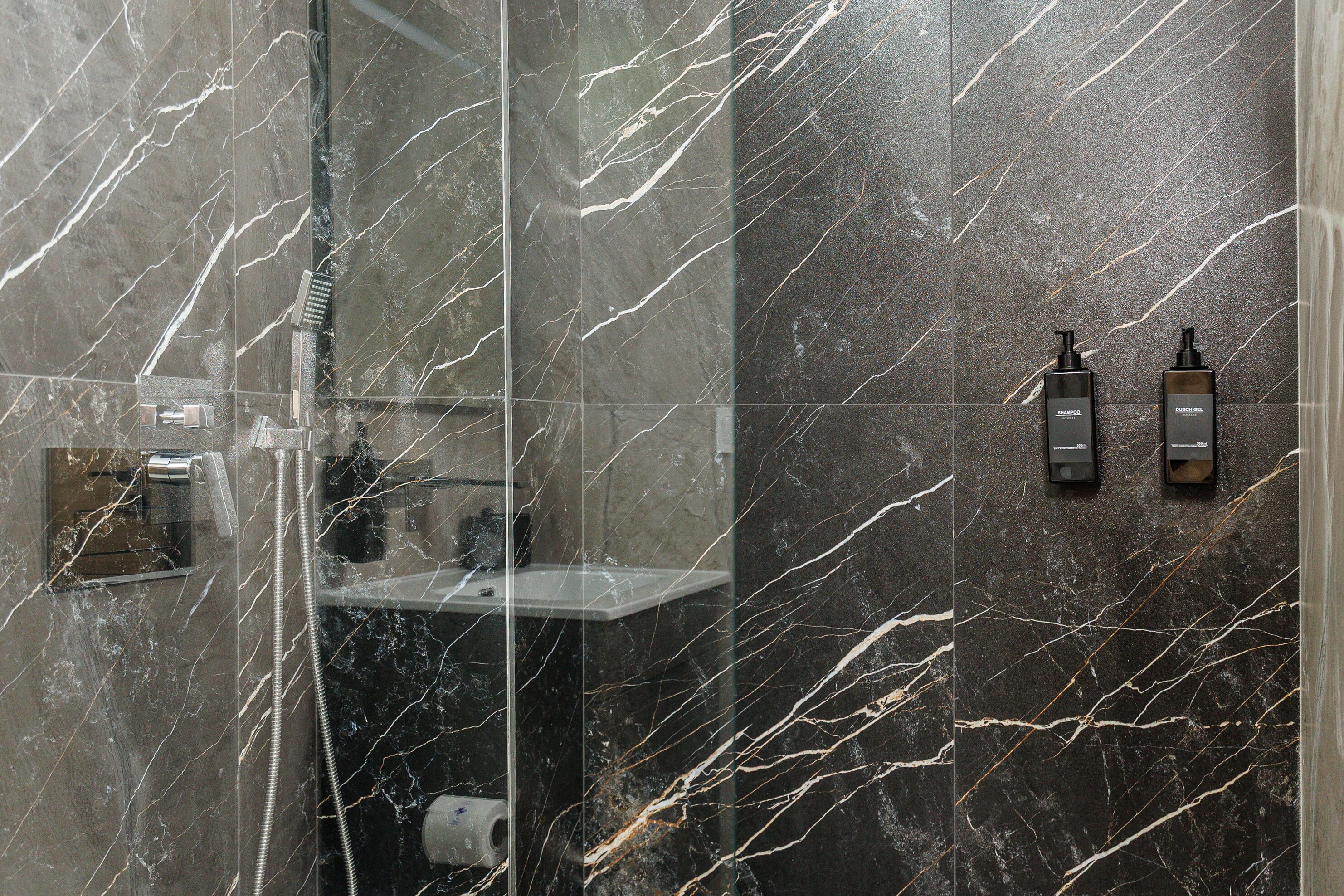 A bathroom with a glass-enclosed shower, a marble wall, and two toiletries on the wall.