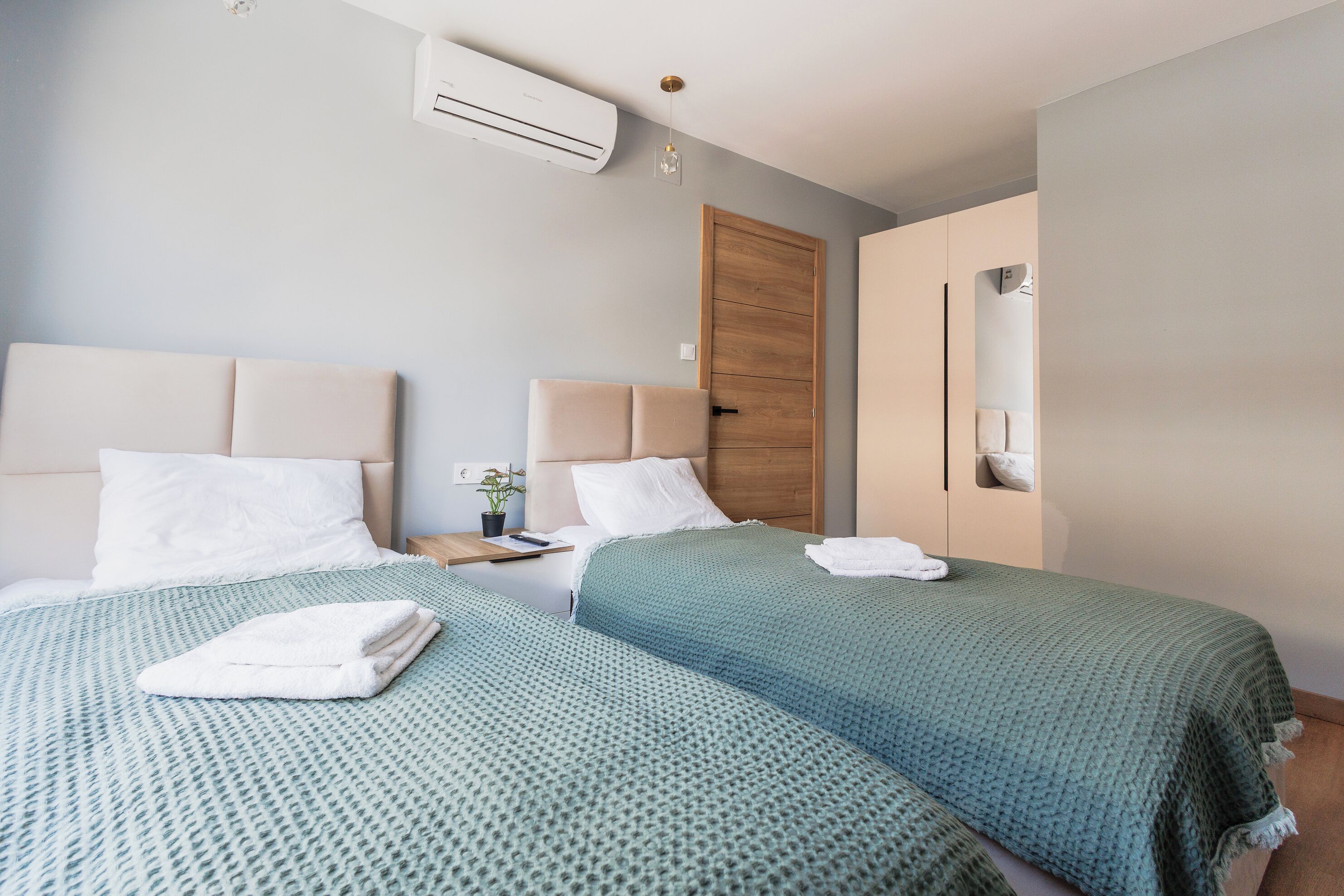 A hotel room with two beds, a wooden door, an air conditioning unit, a bedside table with a lamp, and a small potted plant.