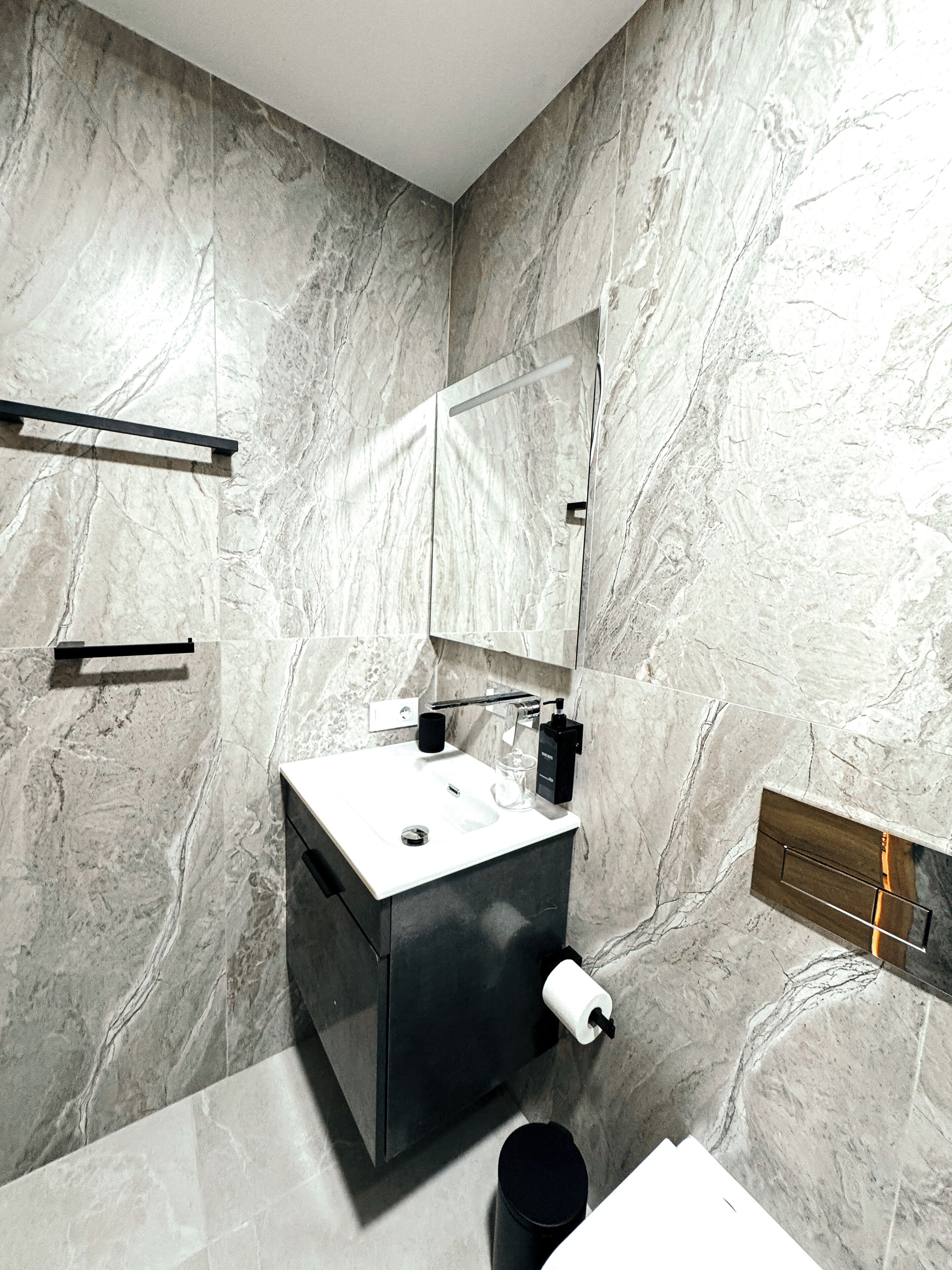 A bathroom with a marble wall, a sink, a mirror, a towel rack, and a toilet paper holder.