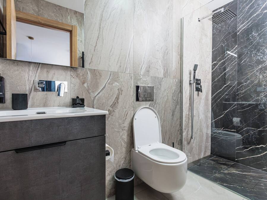 Un baño moderno con pared de mármol, inodoro blanco, lavabo con dispensador de jabón negro y zona de ducha.