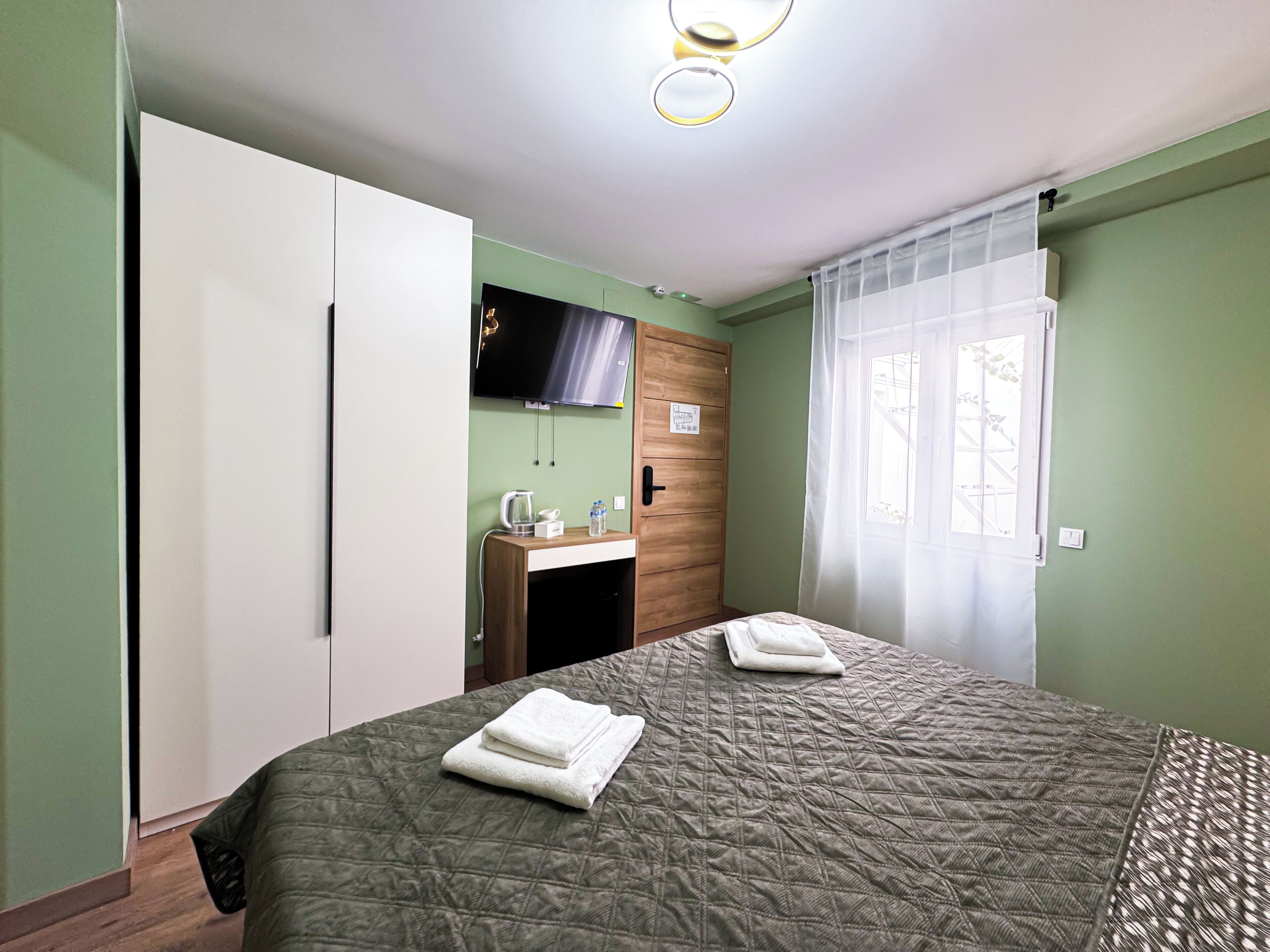 A bedroom with a bed, a wooden wardrobe, a desk with a chair, a television, and a window with curtains.