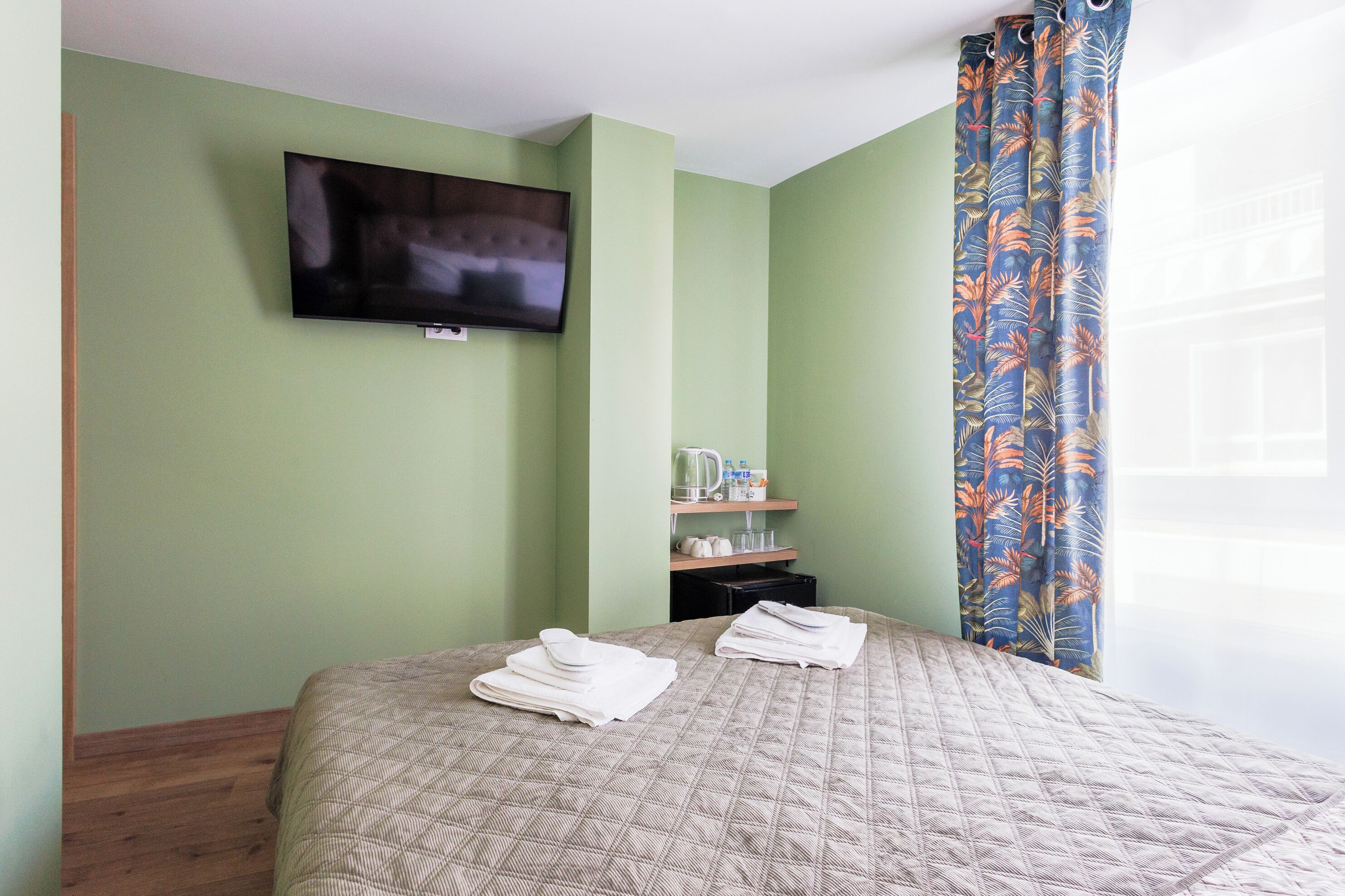 A hotel room with a bed, a television mounted on the wall, a window with a curtain, and a small shelf with items.