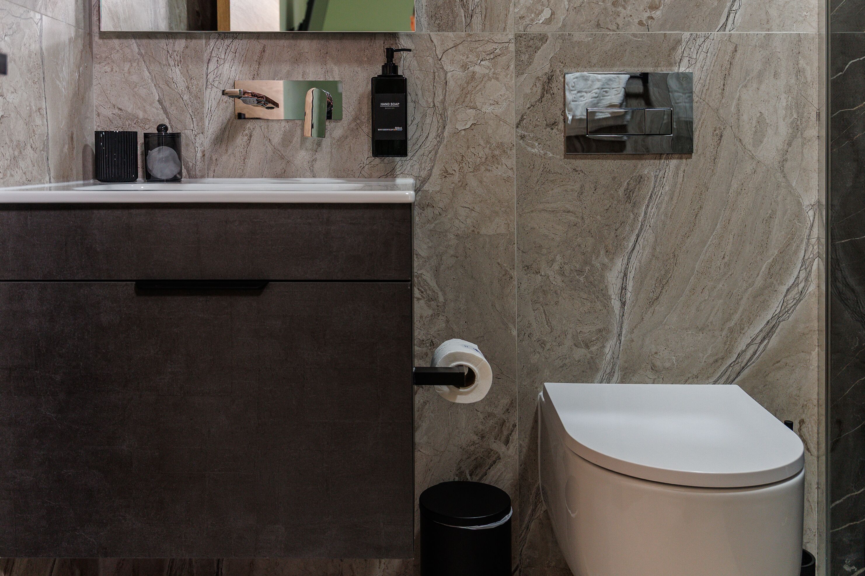 Un baño moderno con pared de mármol, un inodoro, un lavamanos con dispensador de jabón y un cesto de basura.