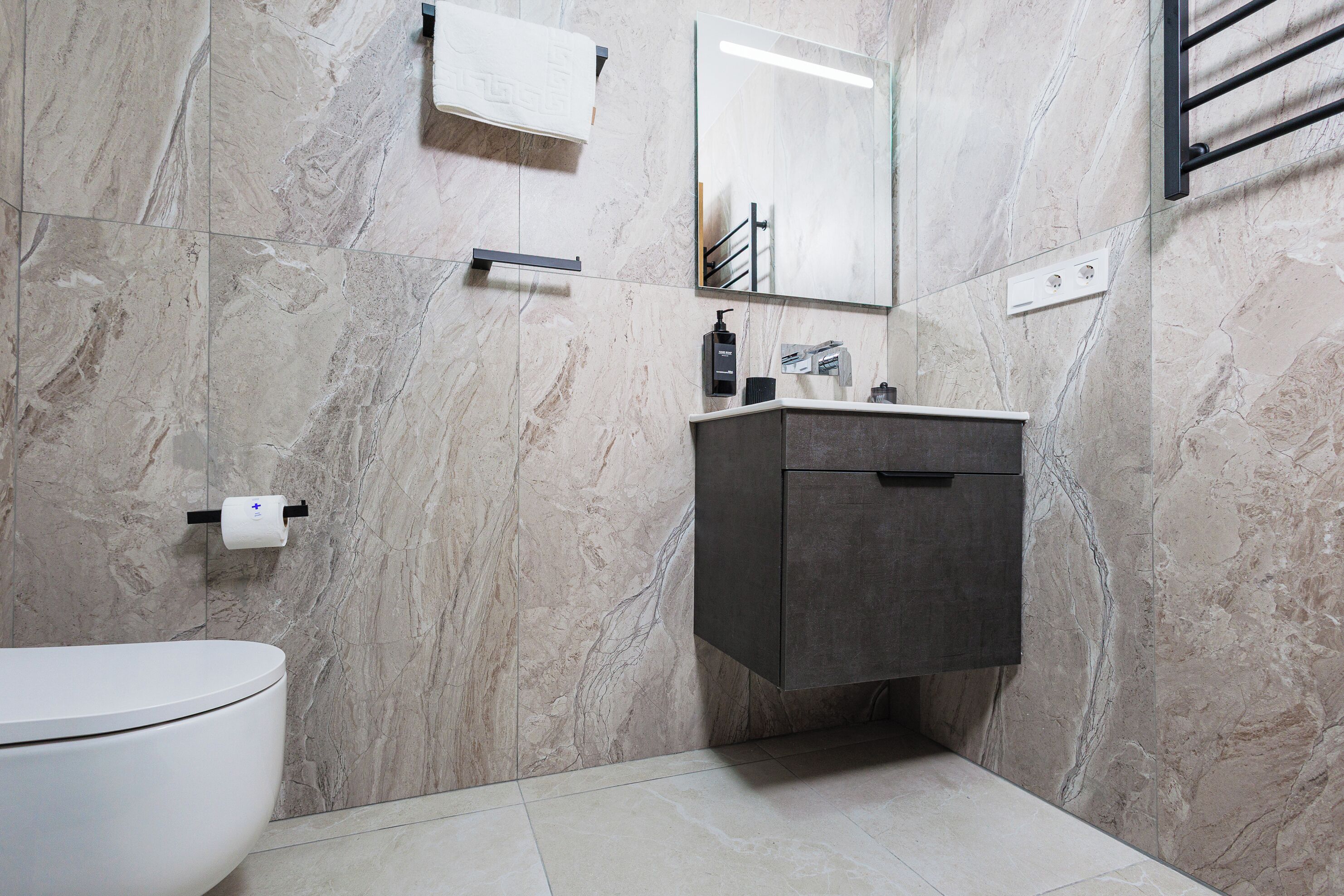 Une salle de bain moderne avec un mur en marbre, des toilettes, un meuble-lavabo foncé et un miroir.