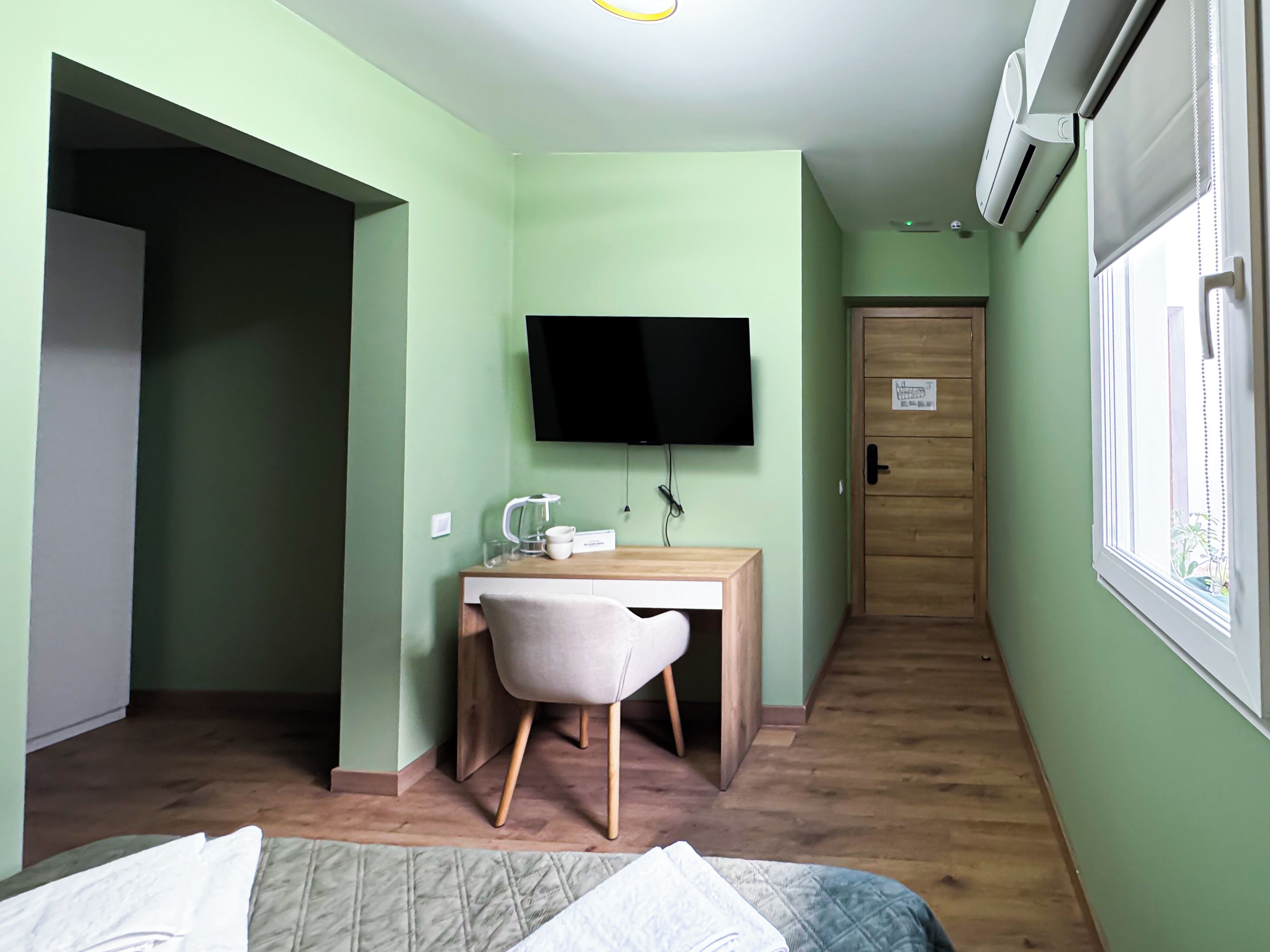 A small, well-lit room with a bed, a desk with a chair, a television, and an air conditioning unit.