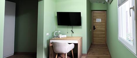 A small, well-lit room with a bed, a desk with a chair, a television, and an air conditioning unit.