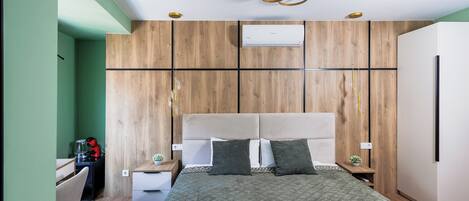 A modern bedroom with a large bed, wooden panel walls, a built-in air conditioner, and a small desk with a chair.