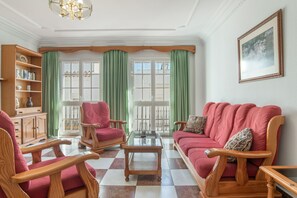 Ein Wohnzimmer mit Schachbrettmuster-Boden, einem roten Sofa, Holzstühlen, einem Glastisch, einem Holzschrank und einem grünen Vorhang.