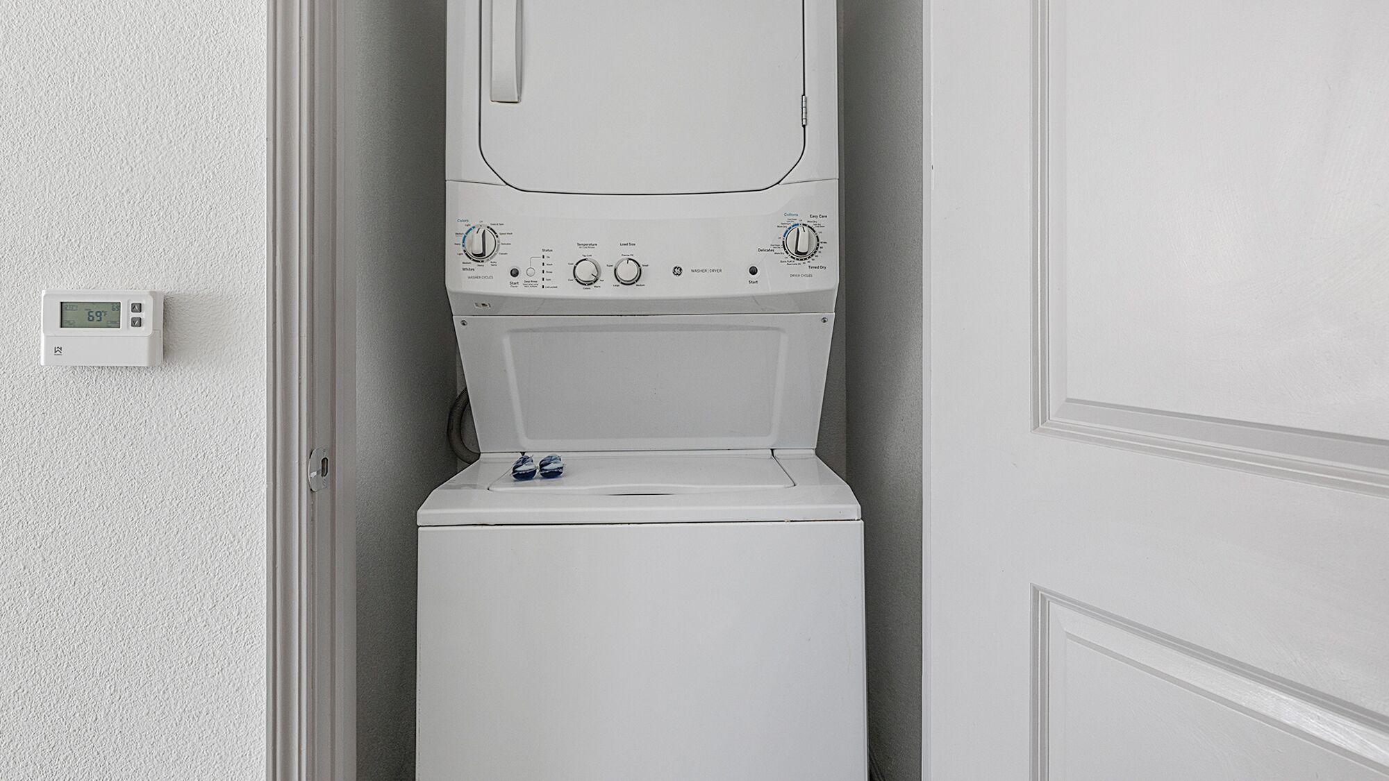 Une machine à laver à chargement frontal, de couleur blanche, se trouve dans une petite buanderie. Un thermostat est fixé au mur.